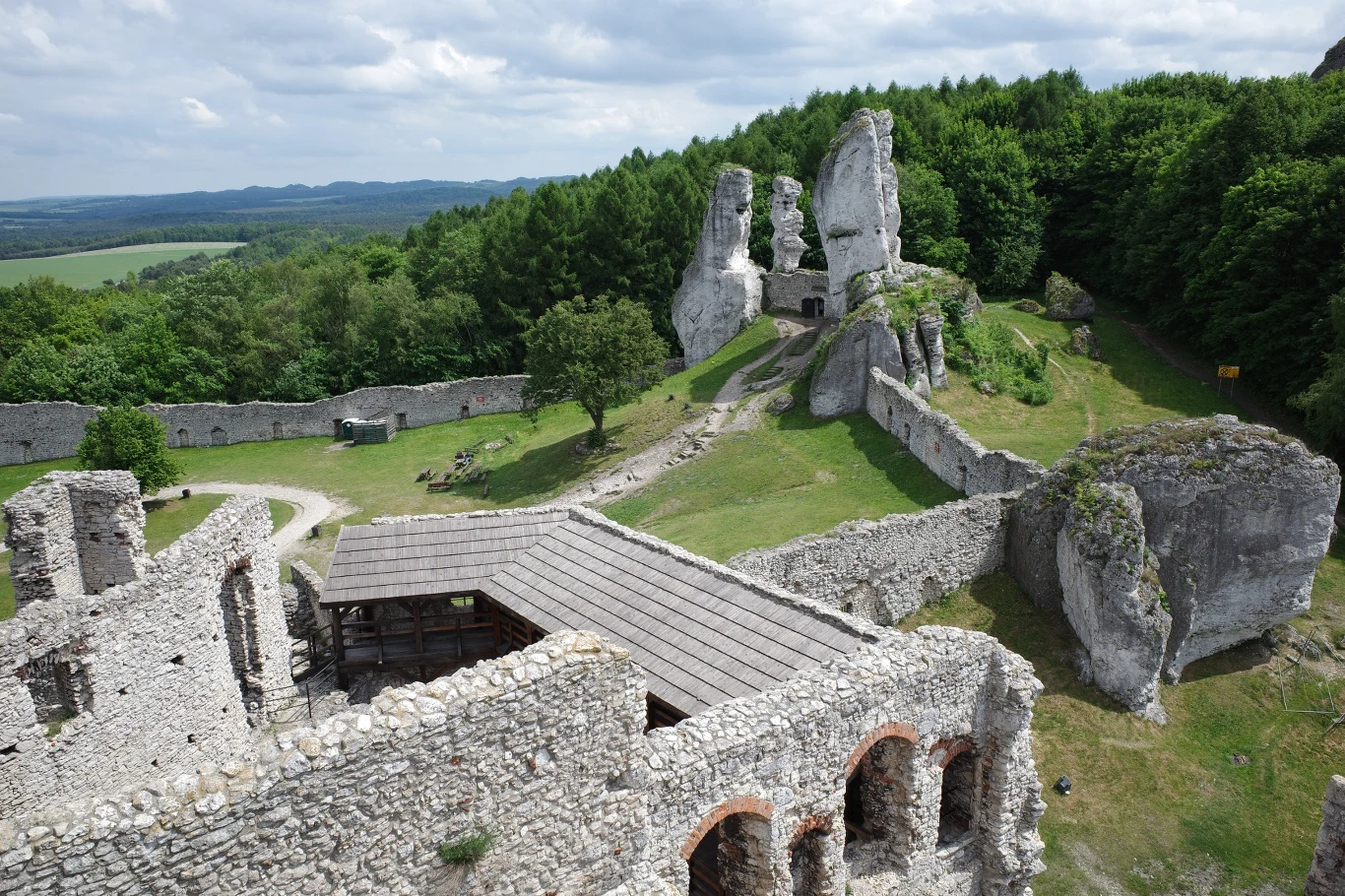 Ruiny Zamku Ogrodzieniec zachwycają położeniem, monumentalnością oraz historią. Kamienne ruiny zamku otoczone zielonym lasem, charakterystyczne wysokie formacje skalne oraz fragmenty murów obronnych na tle rozległego krajobrazu.