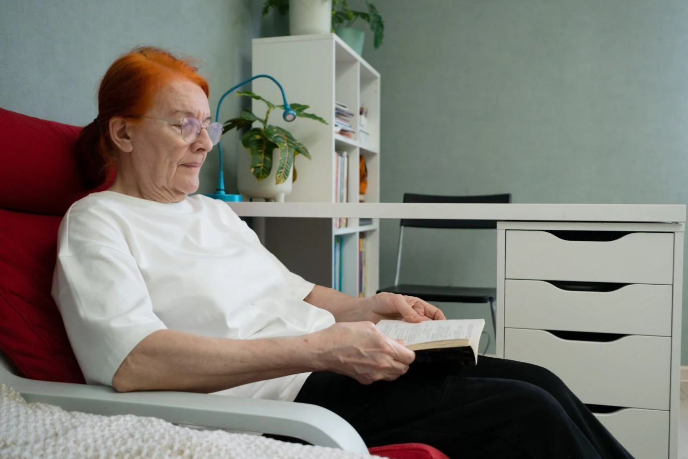 To wspiera pamięć i koncentrację u seniorów. Kluczowy jest codzienny rytm Starsza kobieta z rudymi włosami, ubrana w białą bluzkę i czarne spodnie, siedzi w fotelu i czyta książkę, obok znajduje się białe biurko z szufladami oraz kwiat w doniczce.