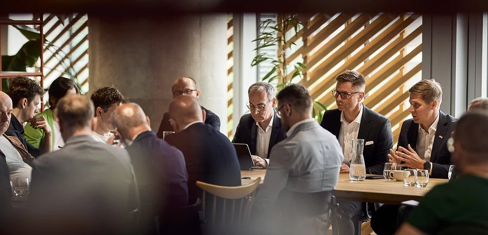 Grupa elegancko ubranych mężczyzn siedzących przy stole w trakcie biznesowego spotkania, z notatnikami i laptopami przed sobą, otoczonych nowoczesną aranżacją wnętrza.