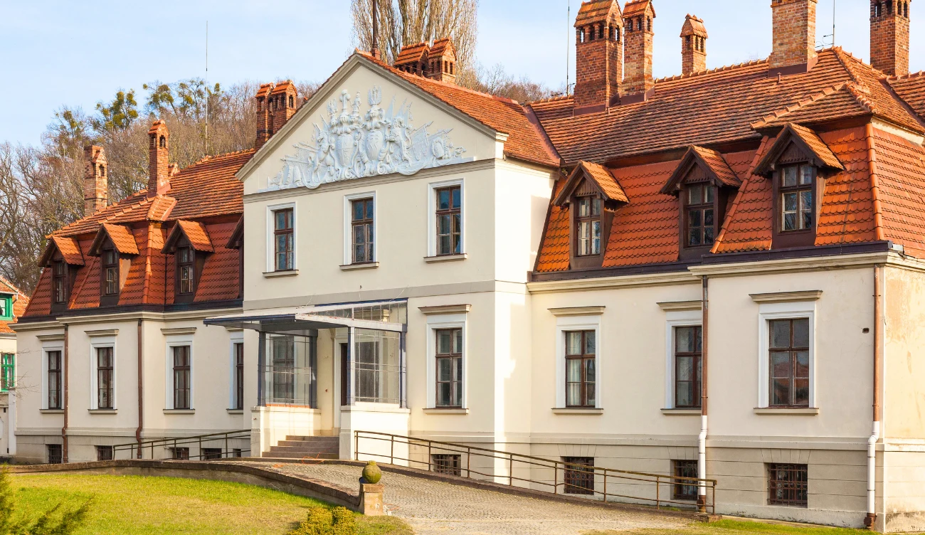 Pałac w Kadynach Zabytkowy budynek o jasnej elewacji z licznymi oknami, spadzistym dachem pokrytym czerwonymi dachówkami i detalami architektonicznymi, w tym ozdobnym tympanonem z płaskorzeźbą.