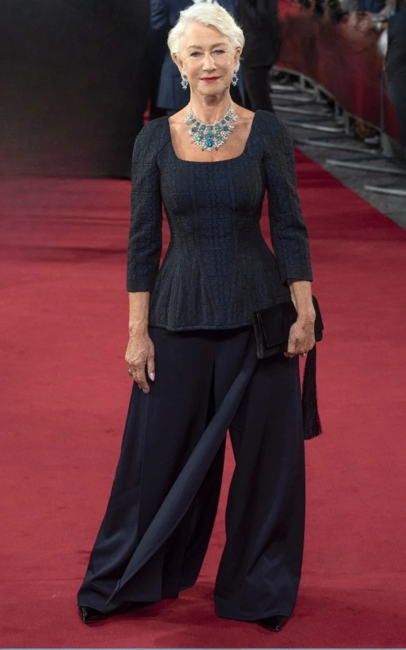 Elegancka kobieta z siwymi włosami ubrana w dopasowaną ciemnogranatową bluzkę, szerokie spodnie oraz wyrazisty, niebieski naszyjnik, trzymająca czarną kopertówkę na tle czerwonego dywanu.