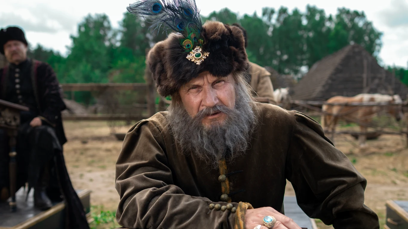 Głównym bohaterem serialu "1670" jest Jan Paweł Adamczewski (w tej roli Bartłomiej Topa) Starszy mężczyzna z długą brodą, ubrany w bogaty strój historyczny z futrzaną czapą zdobioną piórem i broszą, patrzy poważnie przed siebie na tle drewnianych chat i przyrody.