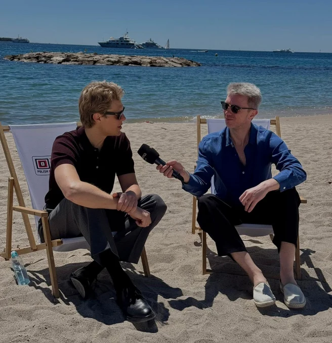 Dwóch mężczyzn siedzi na leżakach na piaszczystej plaży, jeden z nich trzyma mikrofon, przeprowadzając wywiad z drugim; w tle spokojne morze, jachty oraz kamienny falochron.