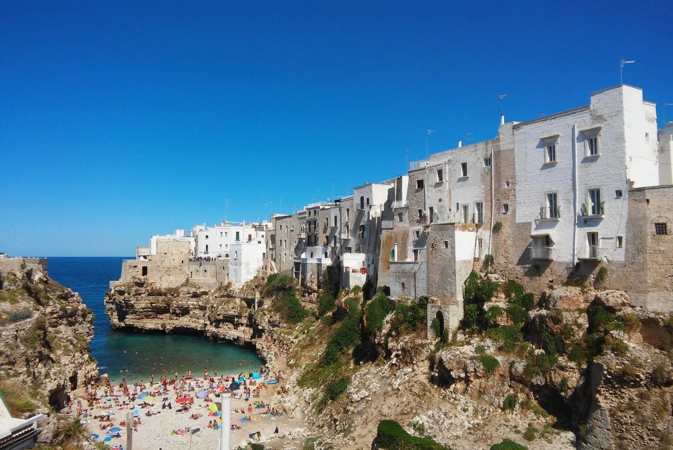 Polignano a Mare to świetne miejsce na jednodniową wycieczkę Plaża otoczona skalistymi klifami, z tłumem ludzi odpoczywających przy morzu i białymi budynkami wznoszącymi się nad zatoką.
