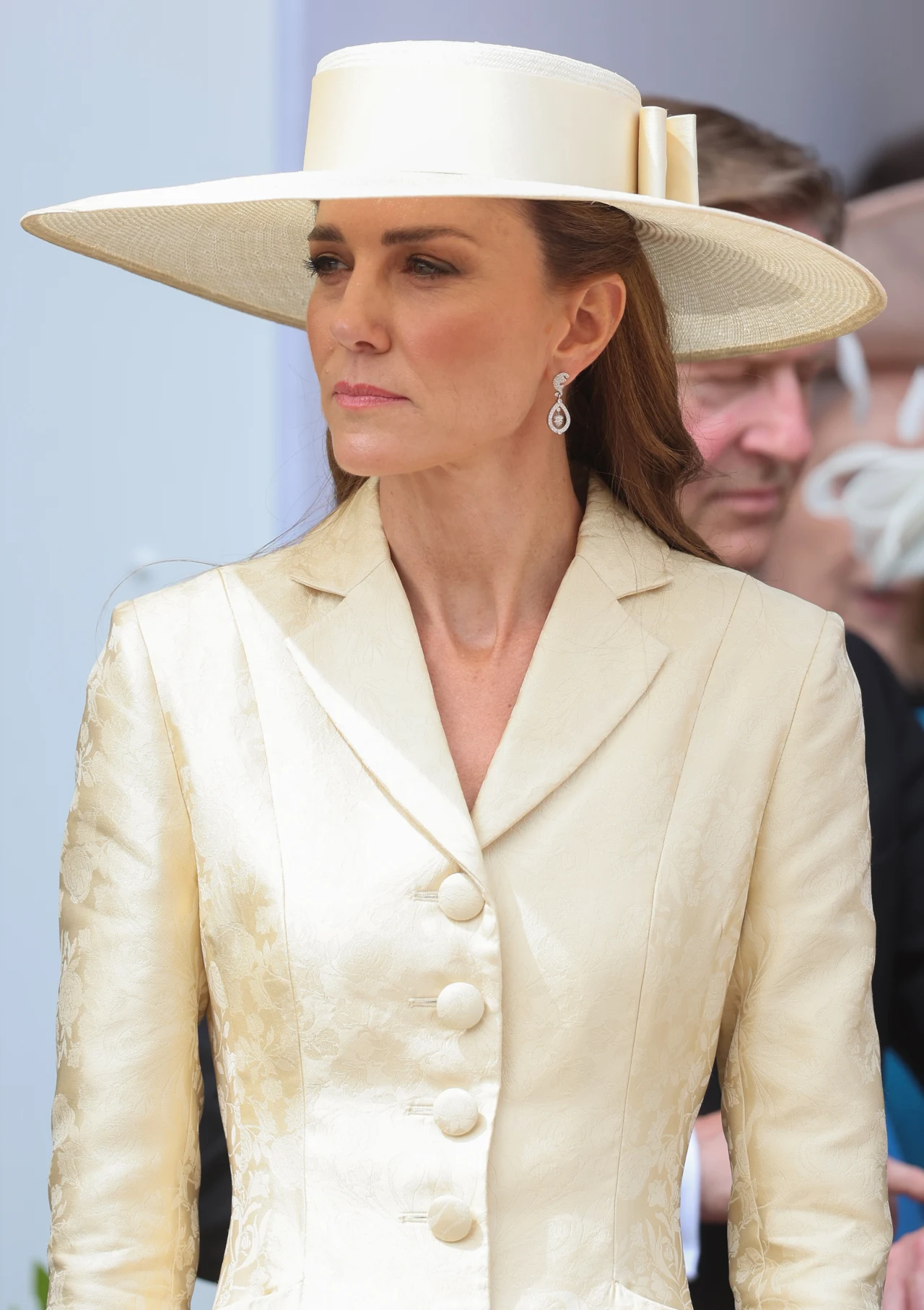 Kobieta w eleganckim kremowym płaszczu i szerokim kapeluszu w tym samym kolorze, nosząca wiszące kolczyki, patrząca przed siebie z poważnym wyrazem twarzy.