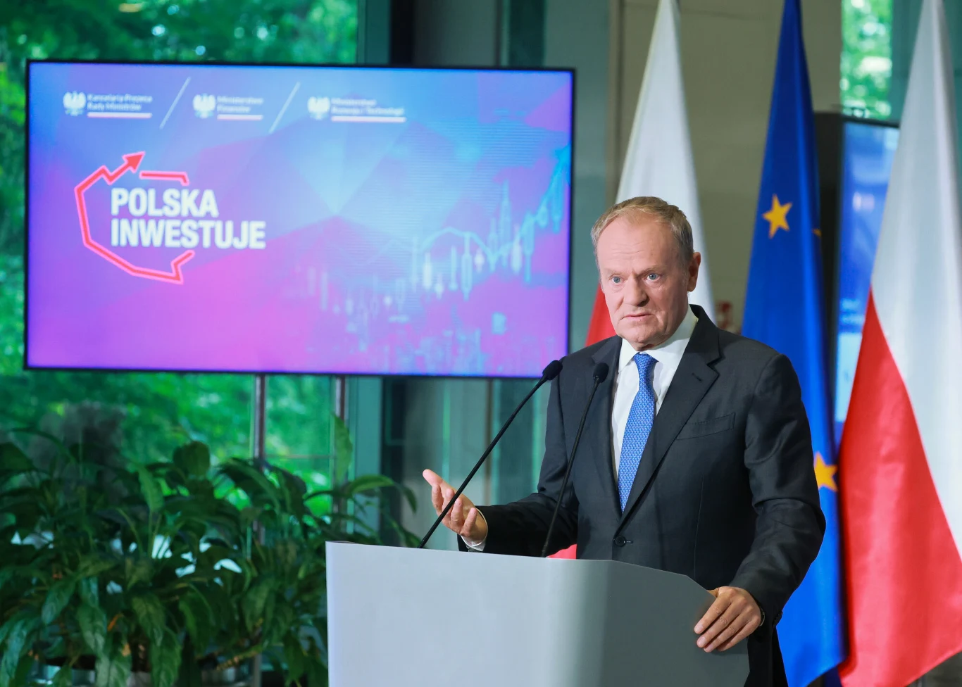 Mężczyzna w garniturze przemawia przy mównicy, w tle ekran z napisem „Polska inwestuje” oraz flagi Polski i Unii Europejskiej.