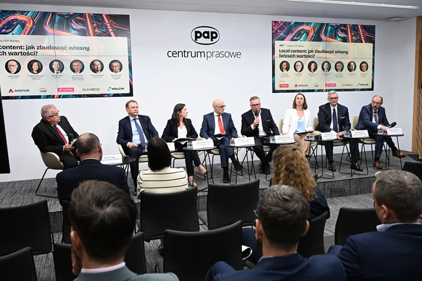 Panel dyskusyjny z udziałem ośmiu prelegentów siedzących przy stole w nowoczesnej sali konferencyjnej, moderowany przez osobę trzymającą mikrofon, z prezentacją tematów na ekranach w tle i uczestnikami słuchającymi wypowiedzi.