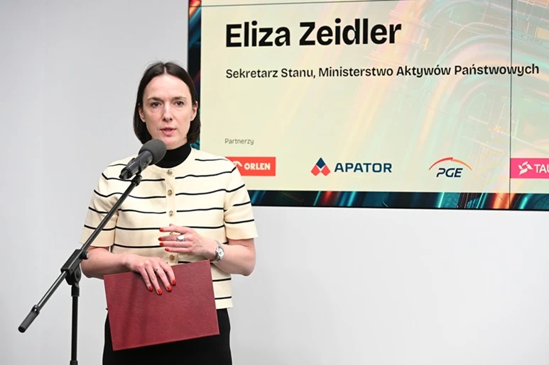 Kobieta w jasnej, paskowanej bluzce przemawia do mikrofonu, trzymając czerwoną teczkę; w tle ekran z napisem 'Eliza Zeidler, Sekretarz Stanu, Ministerstwo Aktywów Państwowych' oraz logotypy firm partnerskich.