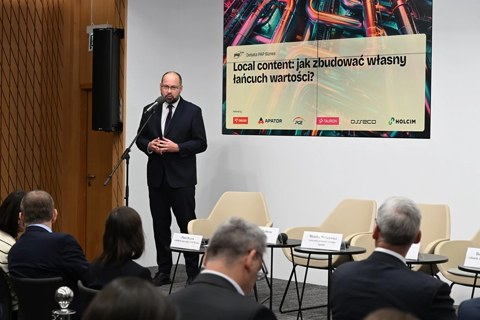 Mężczyzna w garniturze przemawiający przy mikrofonie na tle dużego baneru konferencyjnego z tematem dotyczącym budowania własnego łańcucha wartości, naprzeciwko siedzą uczestnicy wydarzenia, a obok ustawiono puste krzesła i stoliki z nazwiskami prelege...
