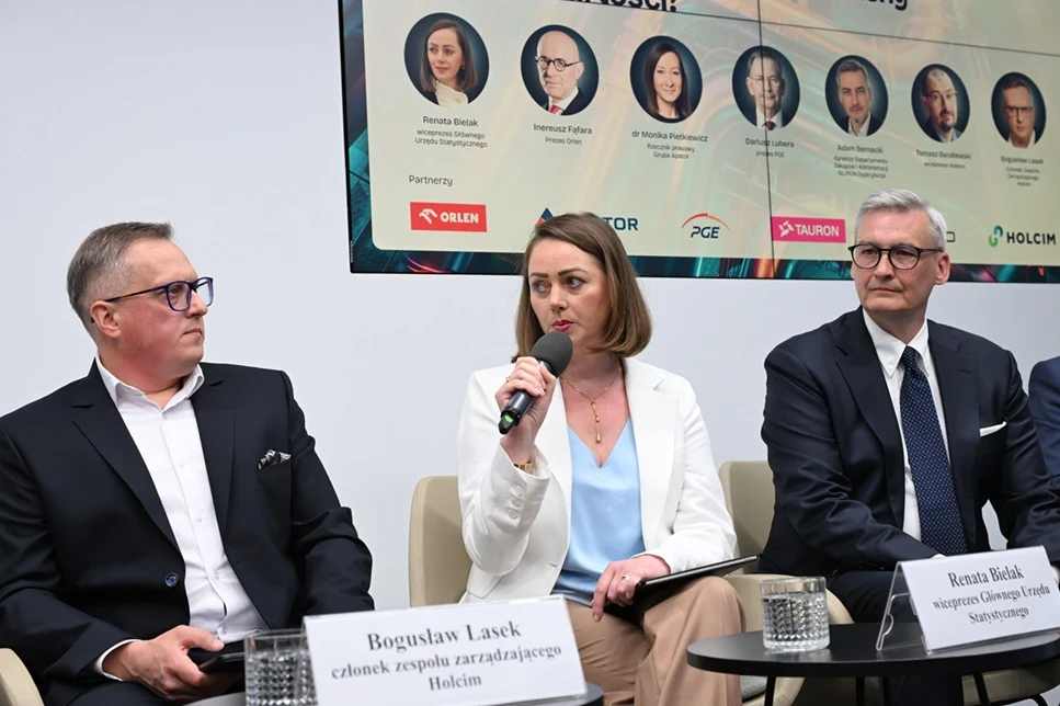 Troje ludzi w formalnych strojach siedzi na panelu dyskusyjnym, kobieta trzyma mikrofon i mówi, obok niej siedzą mężczyźni z notatkami i szklankami na stoliku; za nimi widoczny plakat z informacjami o wydarzeniu i partnerach konferencji.