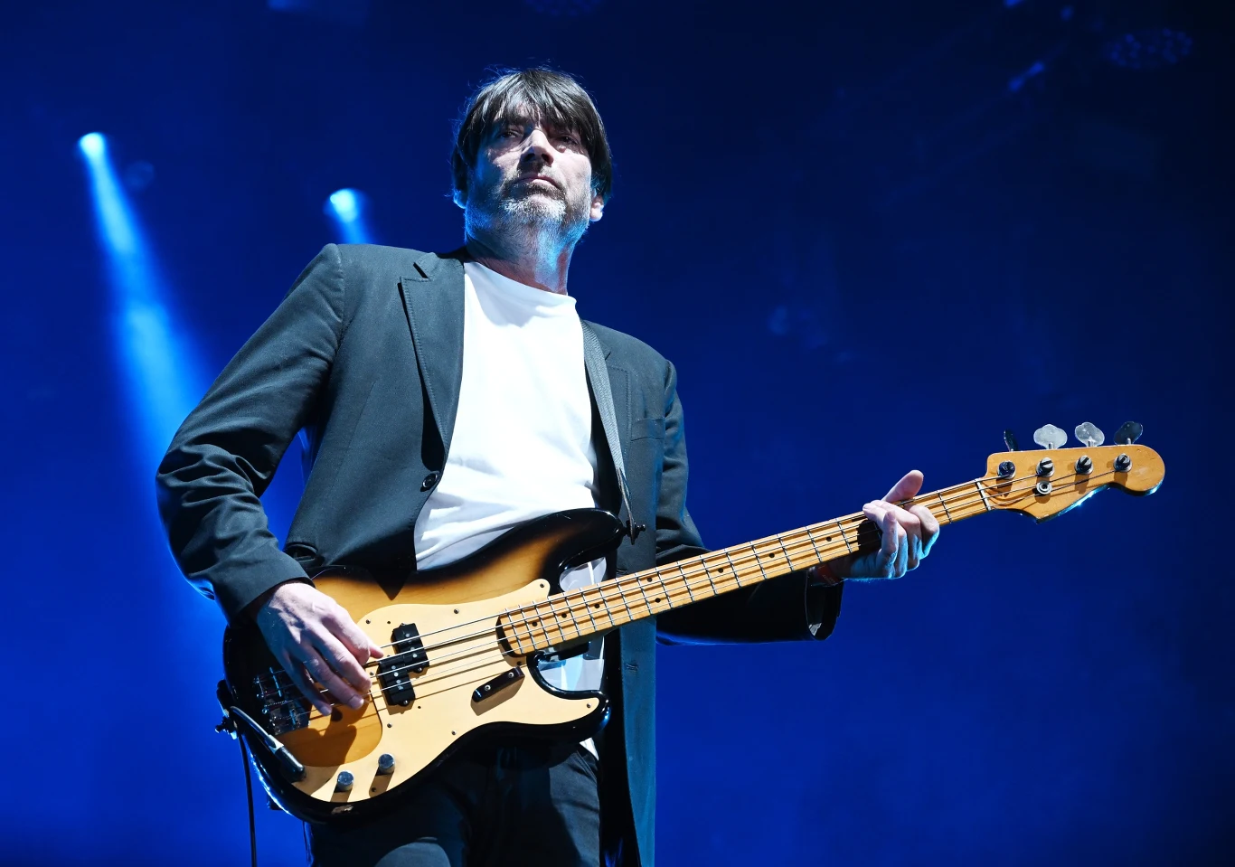 Alex James (Blur) pastiszowym hitem uratował swoje finanse Alex James z Blur w czarnym garniturze gra na gitarze basowej na scenie oświetlonej niebieskim światłem.