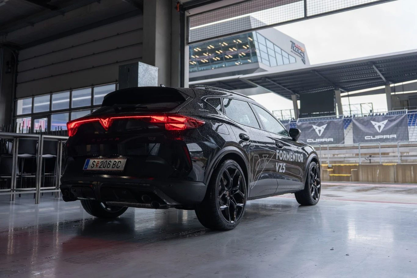 Czarny samochód Cupra Formentor VZ5 z nowoczesnym oświetleniem tylnym zaparkowany w garażu toru wyścigowego, w tle banery reklamowe marki Cupra.