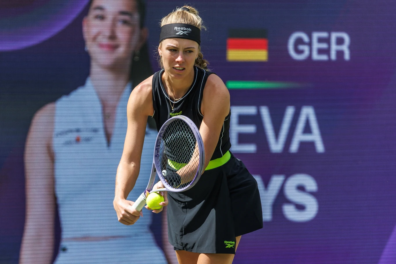 Tenisistka w sportowej czarnej sukience przygotowuje się do serwisu na korcie, trzymając rakietę i piłkę tenisową; w tle duży ekran z flagą Niemiec i napisem GER.