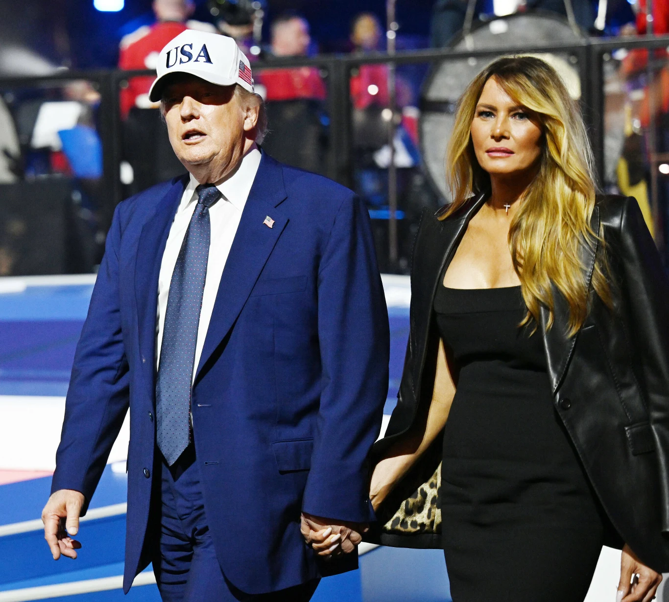 Mężczyzna w granatowym garniturze i białej czapce z napisem „USA” trzyma za rękę kobietę w czarnej sukience i czarnej marynarce z długimi, rozpuszczonymi włosami.