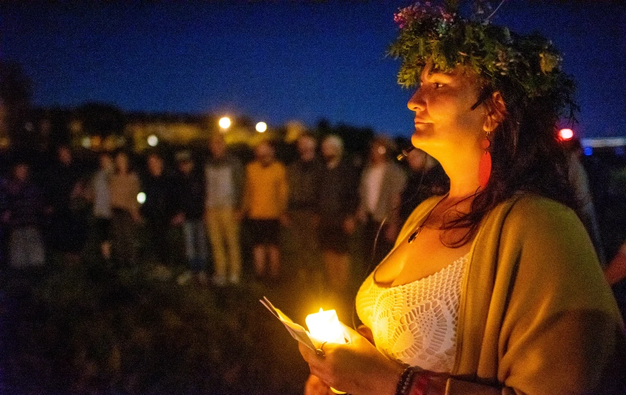 Zwyczaje Nocy Świętojańskiej. Miały zapewnić miłość, pieniądze i zdrowie Kobieta w wianku z liści i kwiatów trzyma świecę, wokół ludzie stojący w kręgu podczas Nocy Kupały po zmroku.