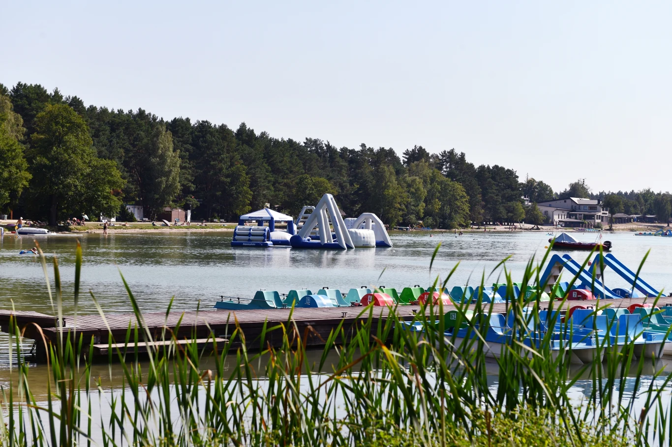 Różnokolorowe rowery wodne ustawione przy drewnianym pomoście, w tle dmuchany park wodny na jeziorze otoczonym lasem.