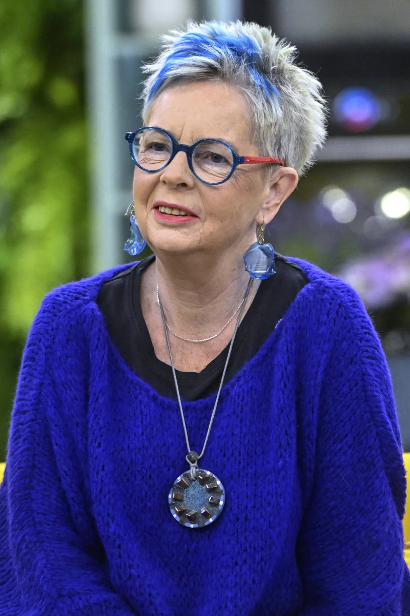 Starsza kobieta z krótkimi siwymi włosami z niebieskimi pasemkami, w okularach i niebieskim swetrze, z dużą srebrną zawieszką na naszyjniku i niebieskimi kolczykami, siedząca w jasnym wnętrzu.