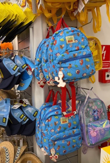 Niebieskie dziecięce plecaki z motywem z bajki Toy Story zawieszone na sklepowym wieszaku obok niebieskich czapek i słomianych kapeluszy.