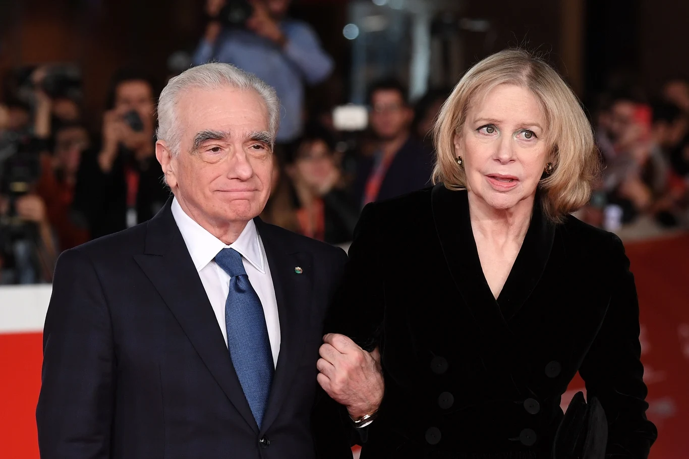 Elegancko ubrany mężczyzna w ciemnym garniturze z niebieskim krawatem idzie pod rękę z kobietą w czarnej aksamitnej sukni z jasnymi włosami, otoczeni przez tłum fotografów podczas oficjalnego wydarzenia.