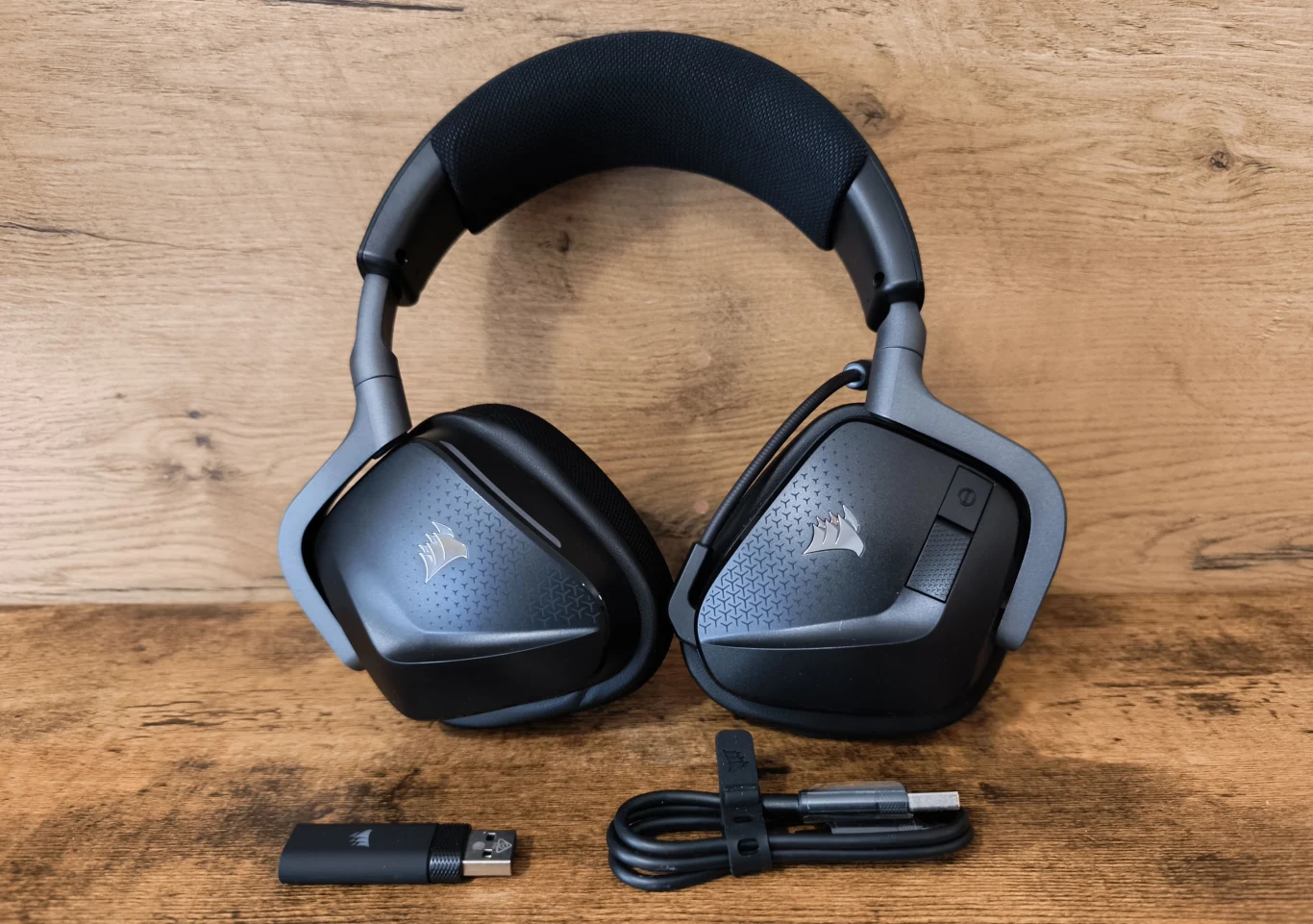 Czarne słuchawki nauszne Corsair z mikrofonem, leżące na drewnianej powierzchni, obok dołączony adapter USB oraz przewód z końcówkami USB-A i USB-C.
