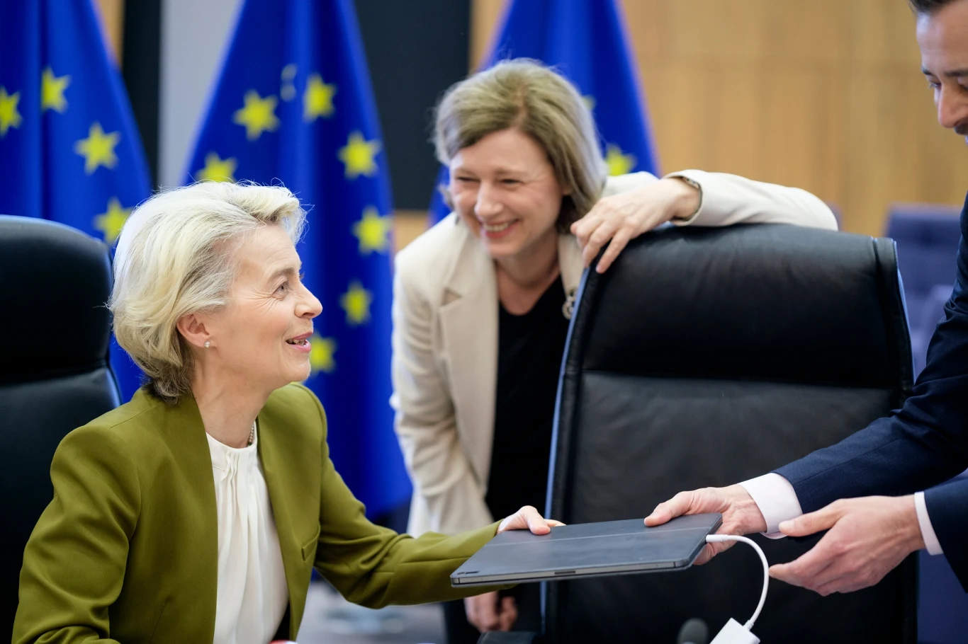 Przewodnicząca Komisji Europejskiej Ursula von der Leyen i była wiceprzewodnicząca KE Vera Jourova Kobieta w zielonej marynarce odbiera dokument od mężczyzny, obok stoi uśmiechnięta kobieta w jasnej marynarce; w tle widoczne flagi Unii Europejskiej.
