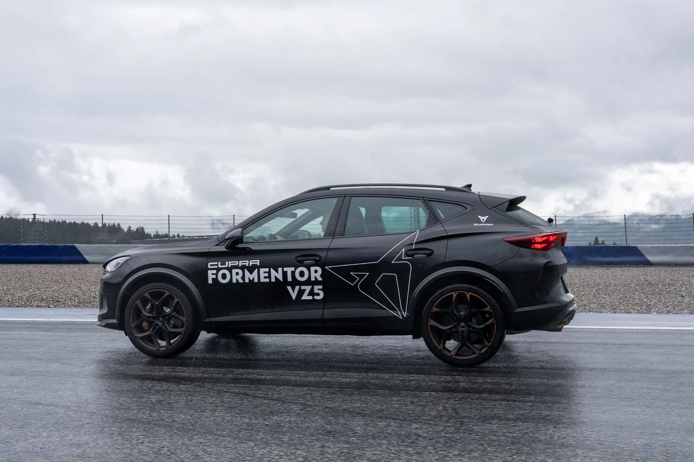 Czarny samochód Cupra Formentor VZ5 z jasnym oznakowaniem na bocznych drzwiach stoi na mokrej nawierzchni toru wyścigowego w pochmurną pogodę.