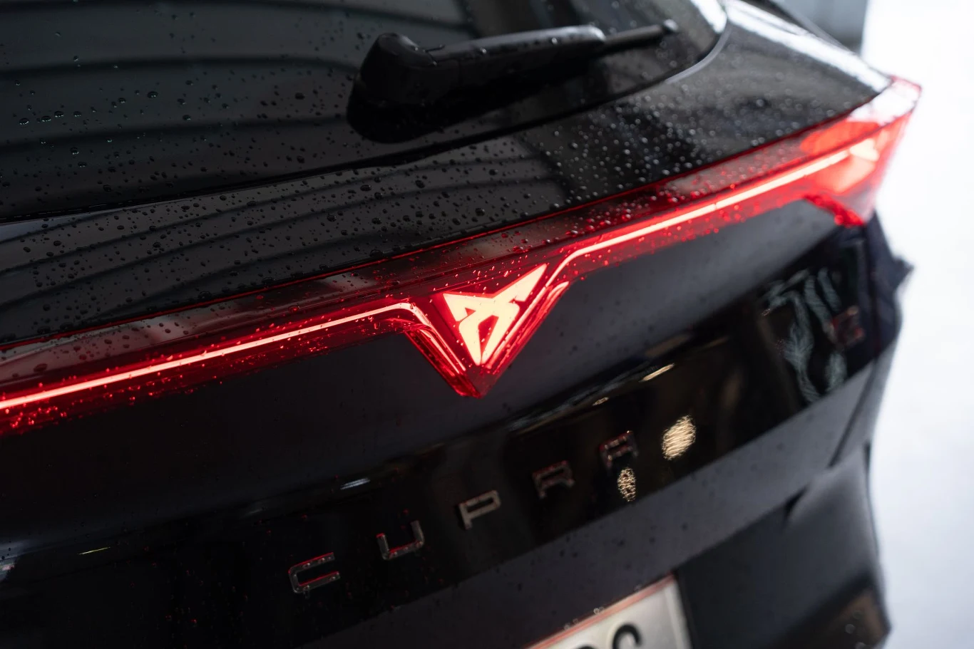 Logo Cupra podświetlone na czerwono na czarnej klapie bagażnika samochodu, z widocznymi kroplami deszczu i refleksami świetlnymi.