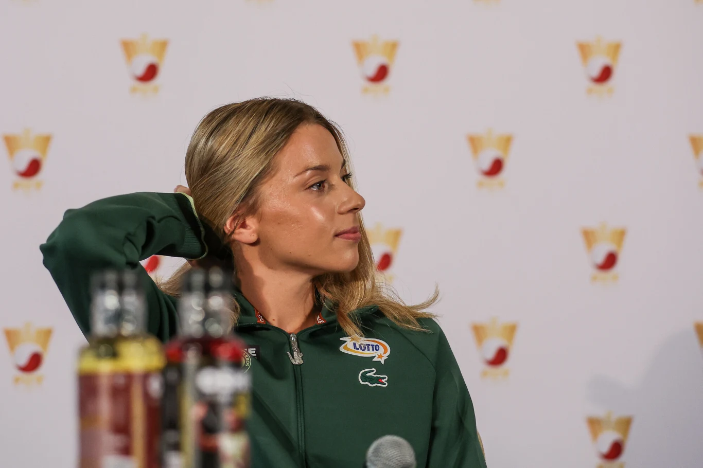 Maja Chwalińska Kobieta w sportowej kurtce z logo Lotto i Lacoste odwraca głowę na bok podczas konferencji prasowej, na pierwszym planie rozmazane butelki i mikrofon, w tle ścianka z logotypami.