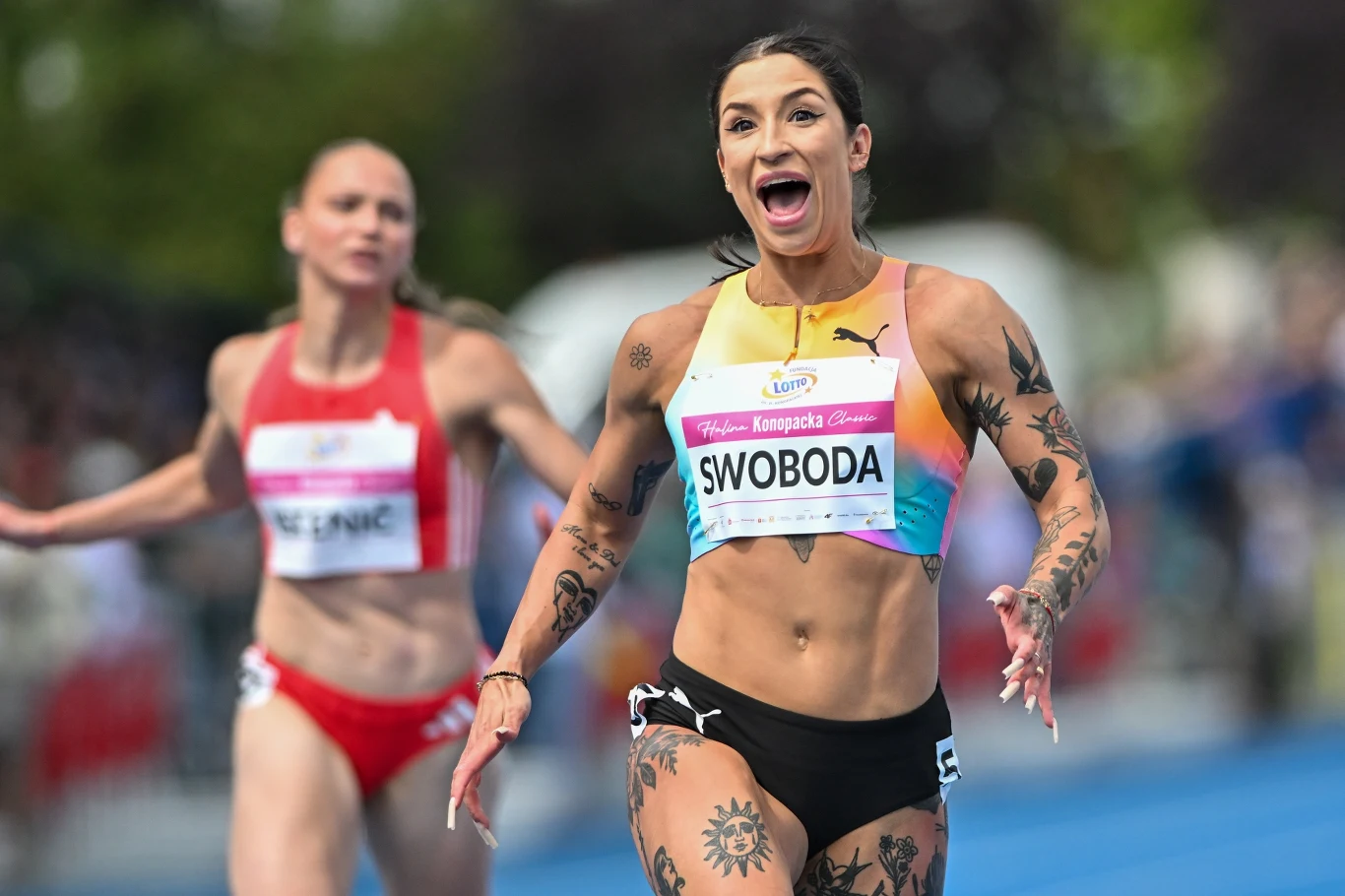 Zawodniczka z widocznymi tatuażami przekracza linię mety podczas wyścigu lekkoatletycznego, wyrażając radość i triumf, podczas gdy rywalka pozostaje z tyłu.