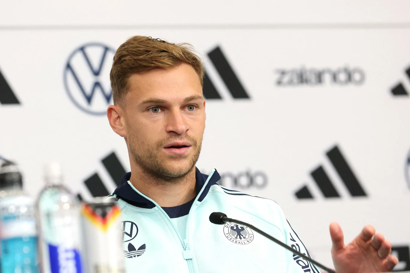 Mężczyzna w sportowej kurtce reprezentacji narodowej siedzi przy stole konferencyjnym, gestykuluje podczas wypowiedzi do mikrofonu, w tle widoczne logotypy sponsorów.