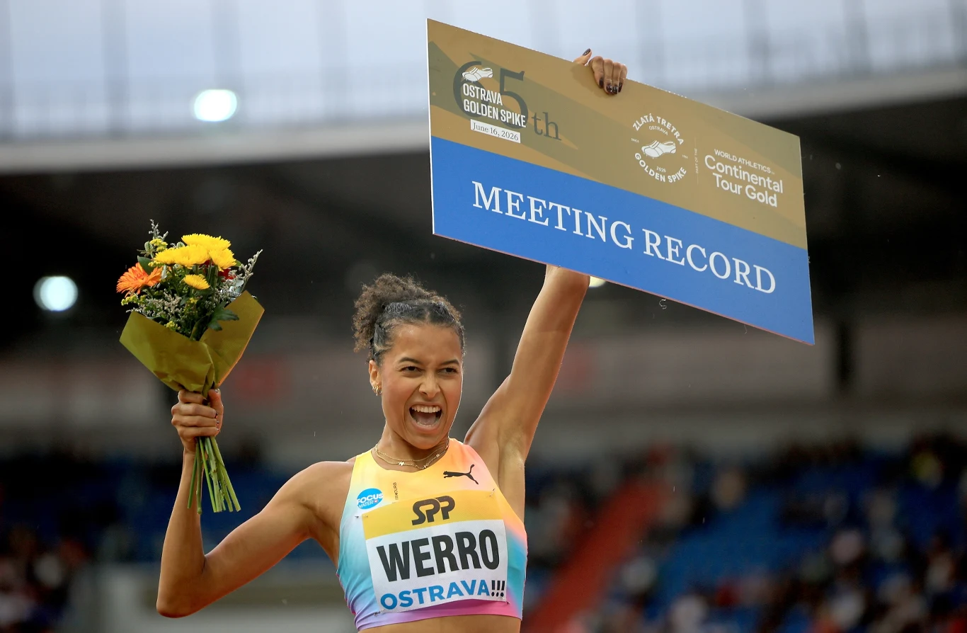 Lekkoatletka w stroju sportowym z napisem „WERRO OSTRAVA!!!” unosi rękę z bukietem żółtych kwiatów oraz dużą tablicę z napisem „MEETING RECORD”, wyrażając radość po ustanowieniu rekordu podczas zawodów na stadionie.