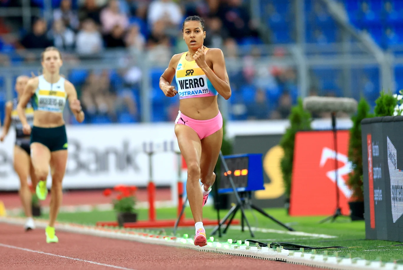 Zawodniczka w różowych spodenkach i żółtym topie biegnie na czele wyścigu na stadionie lekkoatletycznym, wyraźnie skupiona na rywalizacji, tuż za nią pozostałe biegaczki.
