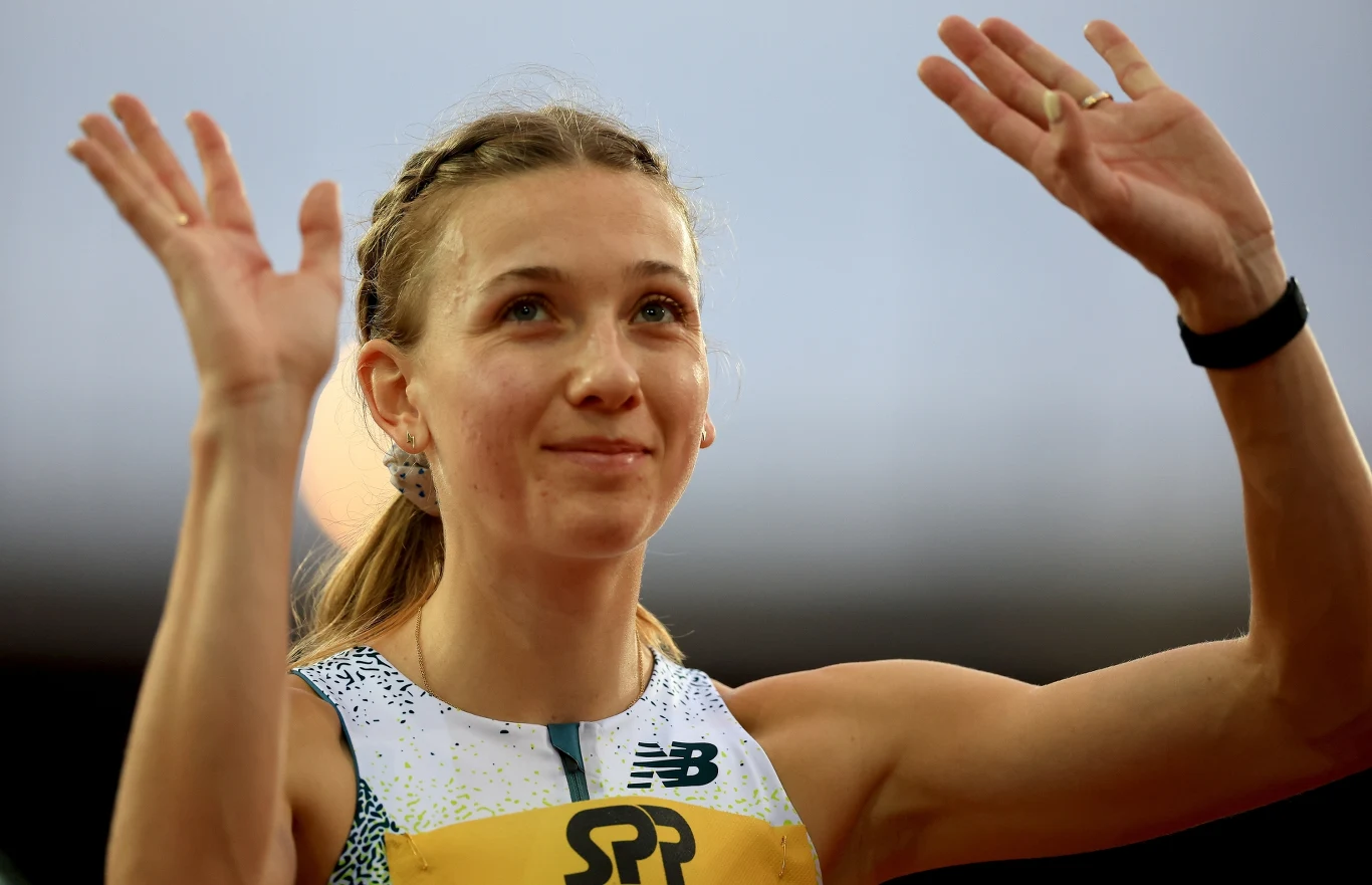 Atletka w sportowym stroju unosi ręce do góry, patrząc z uśmiechem przed siebie.