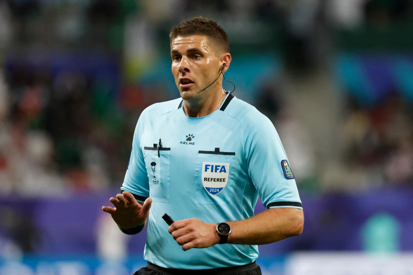 arbiter ubrany w błękitny strój sędziowski z oznaczeniem FIFA, trzymający gwizdek i mikrofon podczas meczu piłkarskiego