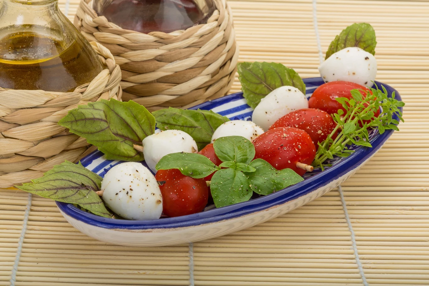 Sałatka caprese z kuleczkami mozzarelli, pomidorkami koktajlowymi oraz świeżymi listkami bazylii, udekorowana pieprzem i podana w owalnym naczyniu, obok butelek oliwy i octu balsamicznego.