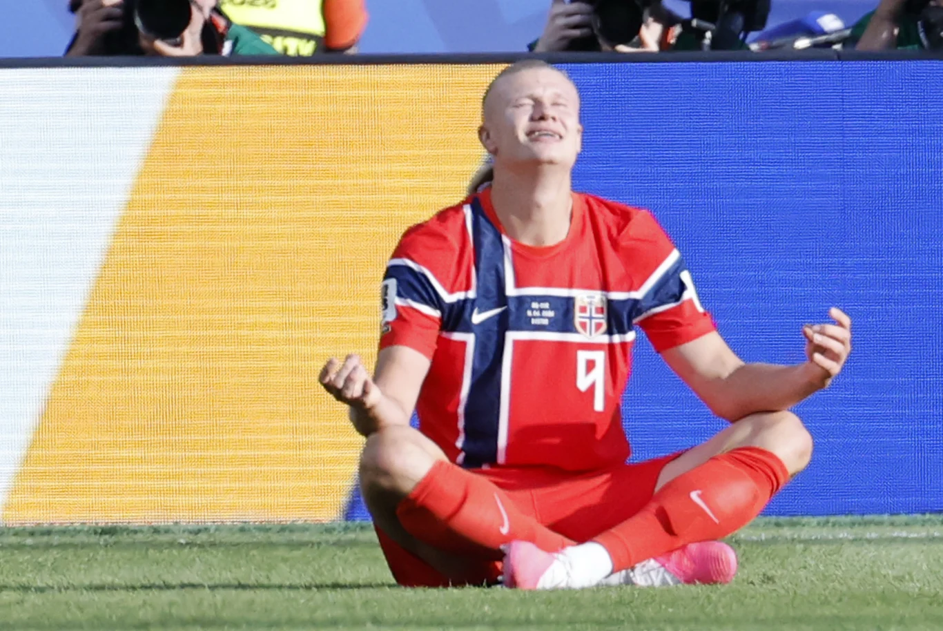 Piłkarz w czerwonym stroju norweskiej reprezentacji siedzi po turecku na murawie z zamkniętymi oczami i uniesionymi dłońmi, wyrażając silne emocje.