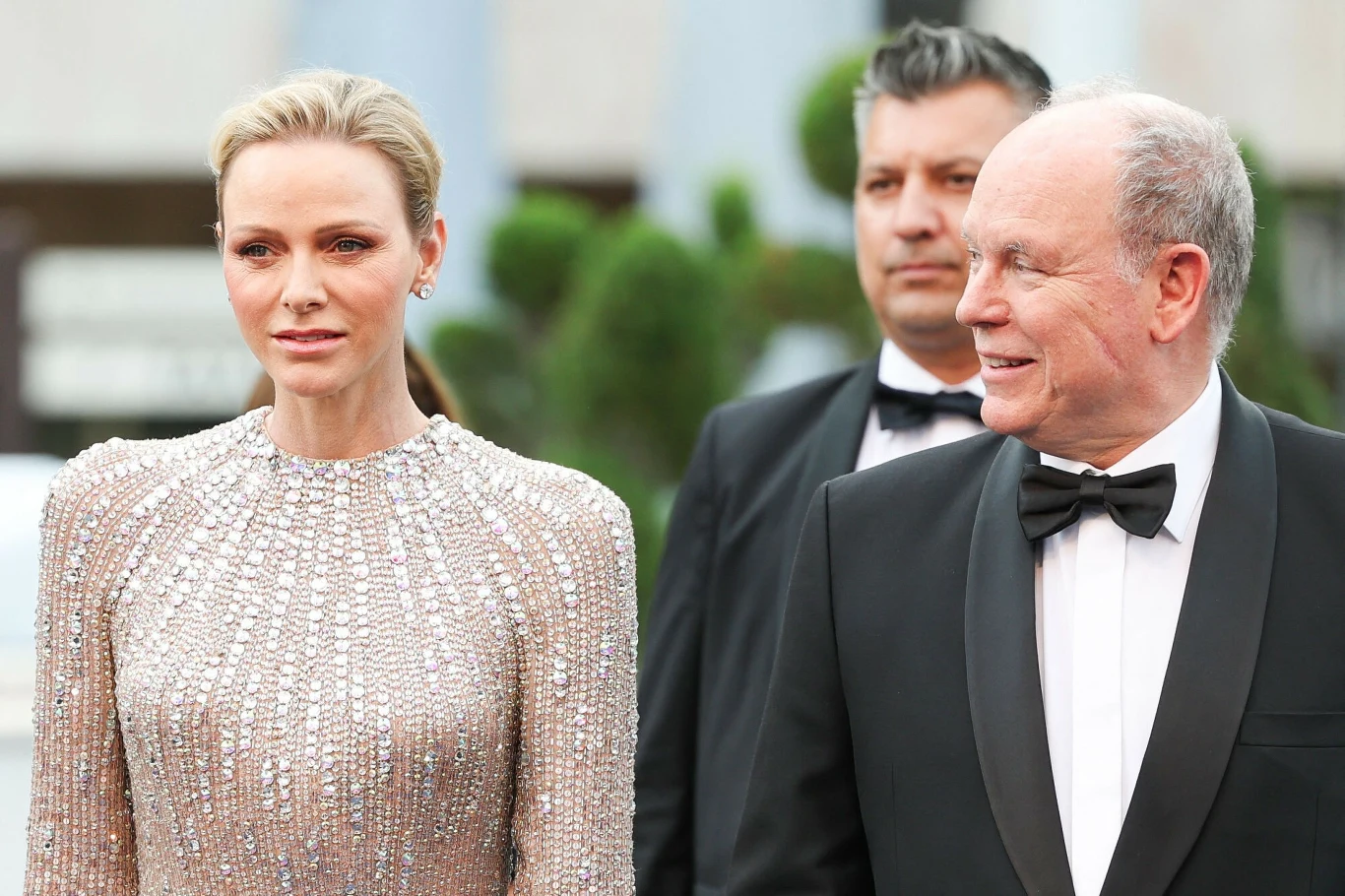 Elegancko ubrani kobieta w błyszczącej, jasnej sukni i mężczyzna w klasycznym smokingu stoją obok siebie, patrząc w różne strony, w tle widoczni kolejni uczestnicy wydarzenia oraz rozmyte elementy otoczenia.
