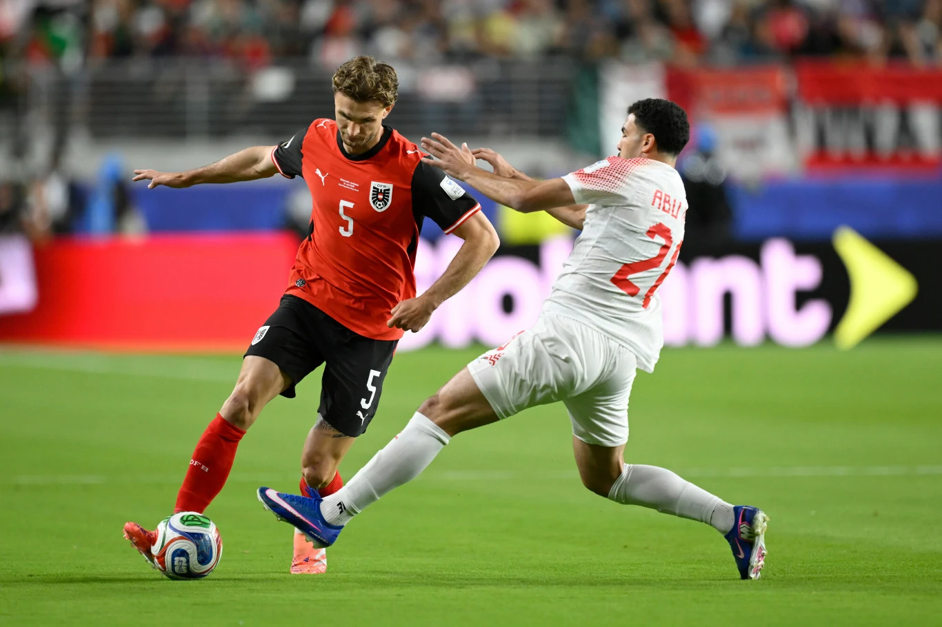 Stefan Posch i Mohannad Abutaha, Austria - Jordania na MŚ 2026 Dwóch piłkarzy rywalizuje o piłkę podczas dynamicznej akcji na boisku; jeden z zawodników ubrany w czerwony strój próbuje przejść obrońcę w białym stroju, który wykonuje wślizg, aby zatrzymać przeciwnika.