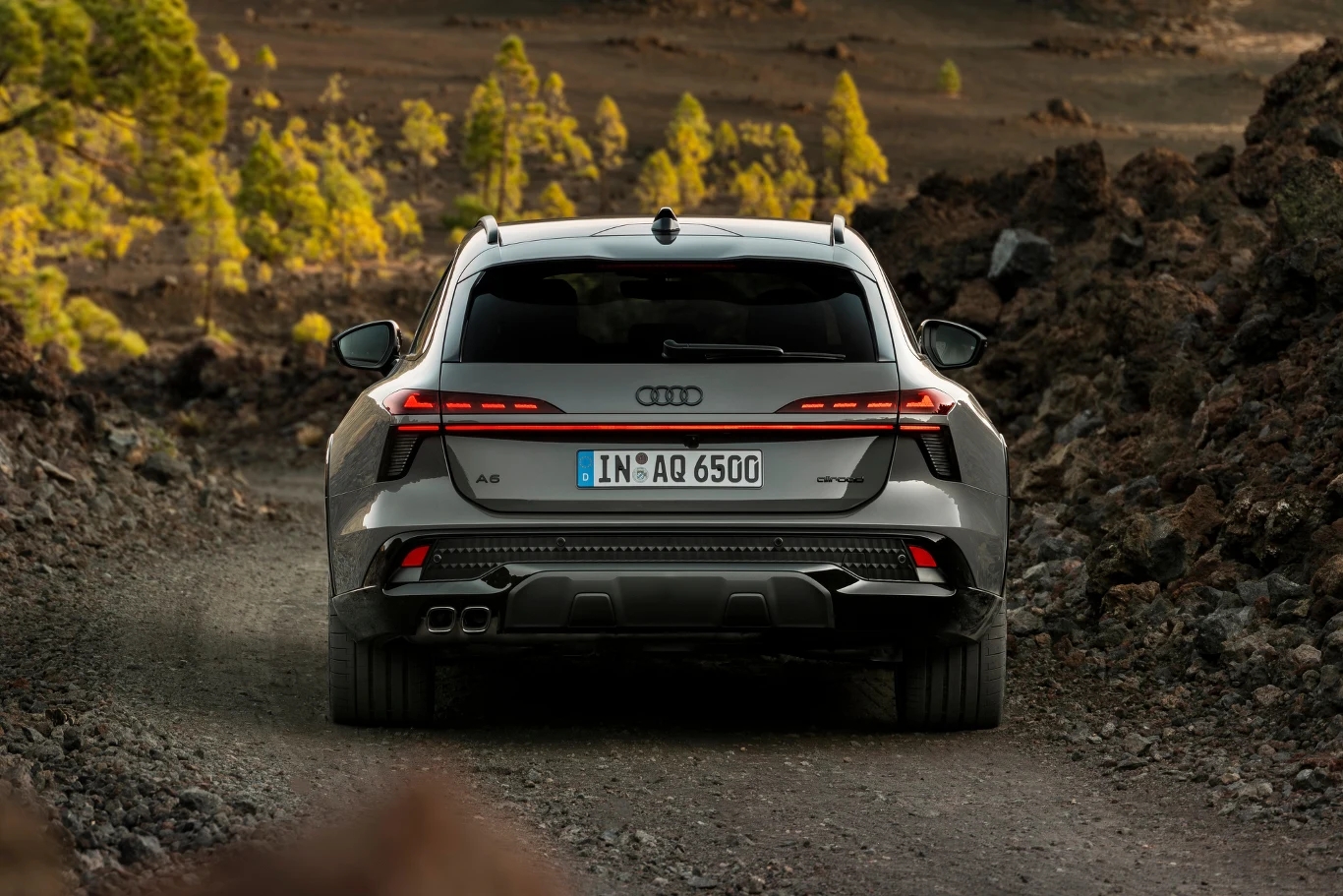 Nadwozie wersji allroad jest szersze o 11 cm względem odmiany Avant. Tył srebrnego Audi A6 z wyraźnymi, nowoczesnymi światłami LED i niemiecką rejestracją, jadącego szutrową drogą przez skalisty teren z widocznymi w oddali zielonymi drzewami.