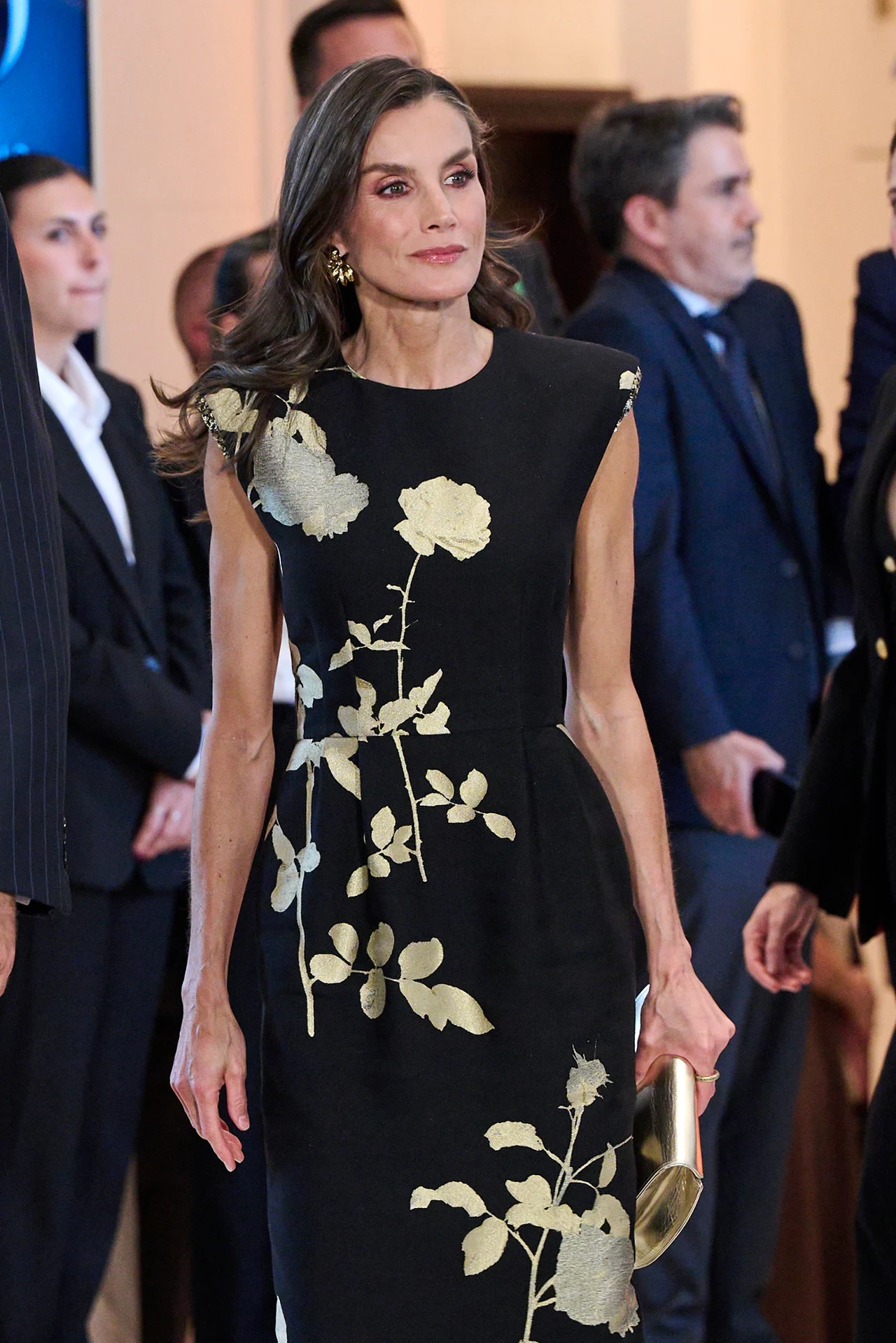Kobieta w eleganckiej czarnej sukni z dużym, kwiatowym wzorem w złotym kolorze, trzymająca złotą kopertówkę, pośród osób ubranych formalnie.