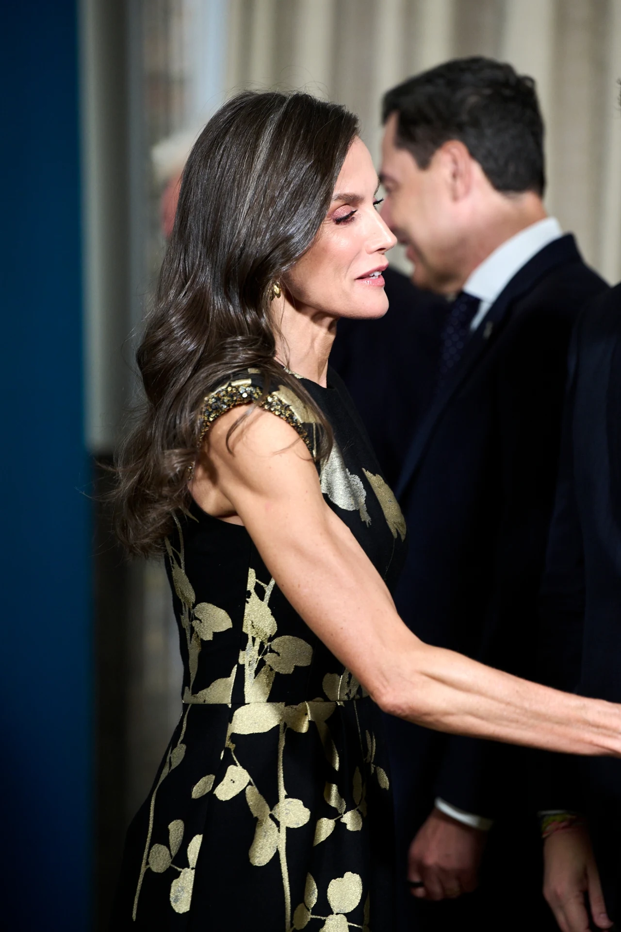Elegancko ubrana kobieta w czarnej sukni ze złotym, kwiatowym wzorem ściska dłoń osobie poza kadrem, w tle widoczni mężczyźni w garniturach.