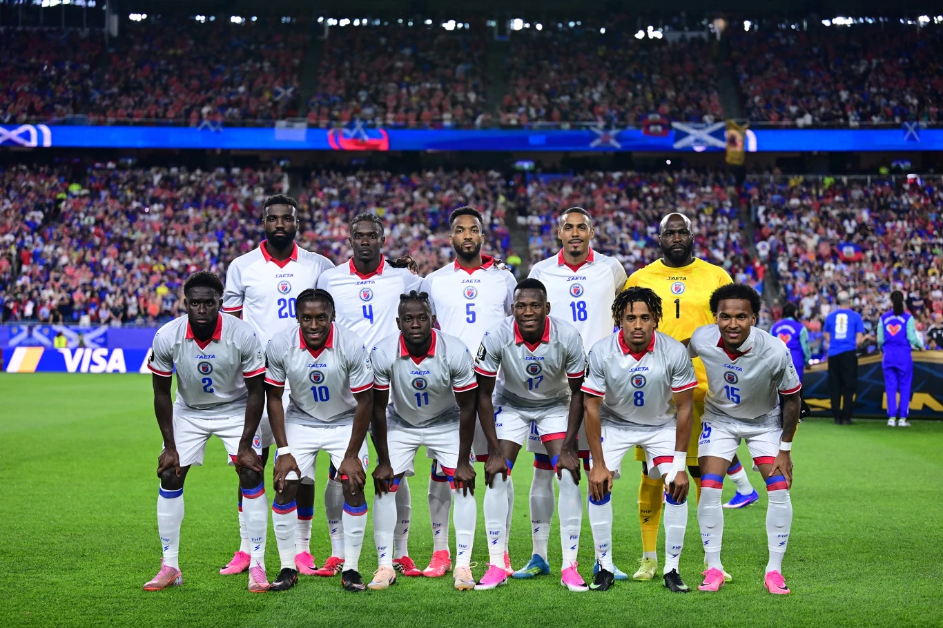 Reprezentacja Haiti w piłce nożnej podczas mundialu 2026 Jedenastu piłkarzy drużyny narodowej w białych strojach pozuje na murawie przed rozpoczęciem meczu, na tle wypełnionego kibicami stadionu.