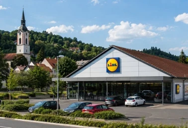 Sklep Lidl z parkingiem w słoneczny dzień, w tle kościół i zalesione wzgórza