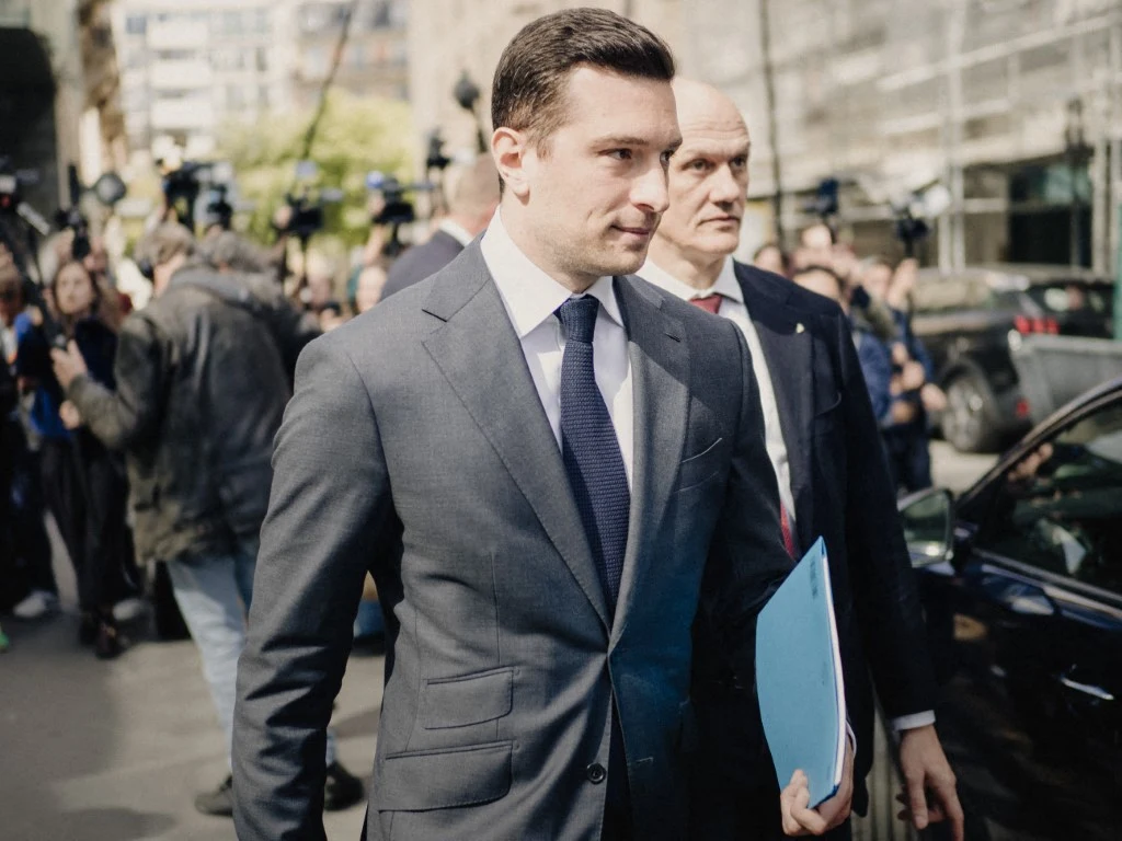 Mężczyzna w eleganckim garniturze, trzymający teczkę z dokumentami, idzie chodnikiem w towarzystwie drugiego mężczyzny, otoczony przez grupę osób i fotografów.