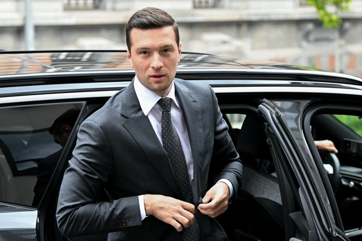 Elegancki mężczyzna w garniturze wychodzi z czarnego samochodu, trzymając nerwowo dłonie przy marynarce, z poważnym wyrazem twarzy.
