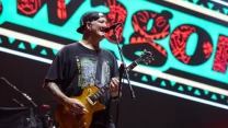 Mężczyzna w czarnej czapce z daszkiem, koszulce z nadrukiem i czarnych spodniach gra na gitarze elektrycznej i śpiewa do mikrofonu na scenie koncertowej, w tle kolorowa grafika z dużymi literami.