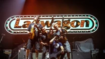 Grupa muzyków z gitarami i mikrofonem pozuje razem na scenie na tle dużego, podświetlanego napisu z nazwą zespołu Lagwagon.