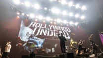 Zespół Hollywood Undead wykonuje energetyczny występ na dużej scenie, mocno oświetlonej reflektorami, z charakterystycznym logo grupy jako tłem ekranowym.