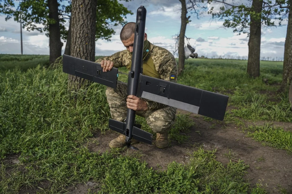 Ukraiński żołnierz z dronem przechwytującym Żołnierz w mundurze kamuflażowym klęczy na ziemi, montując czarny bezzałogowiec o rozpiętych skrzydłach wśród zielonej trawy i drzew.