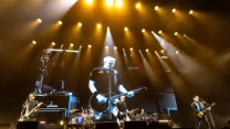 Czterech muzyków gra na dużej scenie podczas koncertu rockowego, za nimi znajduje się ogromny ekran wyświetlający zbliżenie gitarzysty i wokalisty pod jasnym oświetleniem scenicznym.